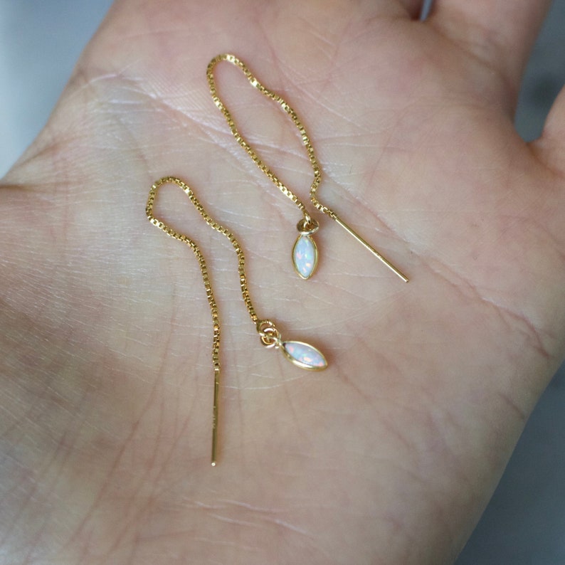 May include: A pair of gold-colored threader earrings with small, oval-shaped opal pendants. The earrings are displayed on a light-colored background, showcasing their delicate design and subtle sparkle. The chain is a fine box chain.