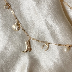May include: A gold chain necklace with multiple charms, including a star, seashell, cowboy boot, horseshoe, and pearl.