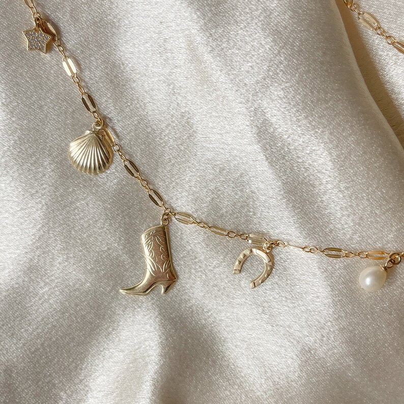 May include: A gold chain necklace with multiple charms, including a star, seashell, cowboy boot, horseshoe, and pearl.