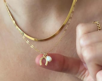 Puede incluir: Un collar de cadena de oro con un delicado dije de herradura y un pequeño colgante de perla. El collar está superpuesto a una cadena de oro más gruesa.