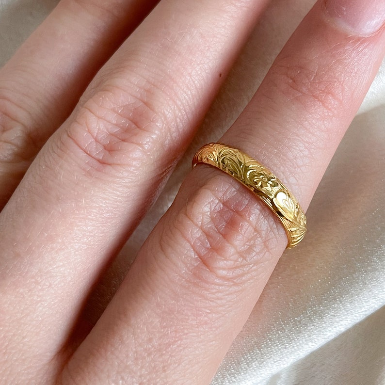 May include: A gold band ring with a floral design. The ring is worn on a finger.