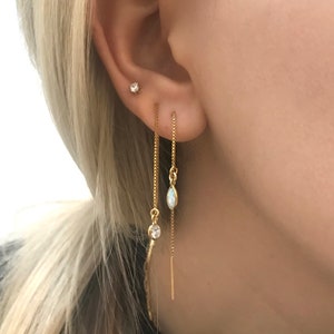May include: Close-up of a person's ear adorned with three gold earrings. One is a small stud, and the other two are dangling chain earrings. One chain earring has a clear stone, and the other has an oval-shaped opal.