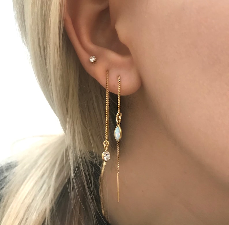 May include: Close-up of a person's ear adorned with three gold earrings. One is a small stud, and the other two are dangling chain earrings. One chain earring has a clear stone, and the other has an oval-shaped opal.