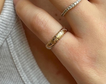 May include: Close-up of a hand wearing three gold rings. The primary ring is a gold band with an engraved floral pattern. A second ring is a thin gold band with small, clear stones. A third ring is a thick gold chain link style.