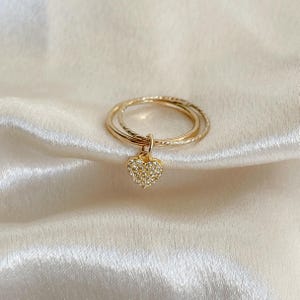 May include: A gold-colored ring set with two bands, one textured, and a dangling heart pendant. The heart is encrusted with small, clear crystals. The ring rests on a white, satin-like fabric.