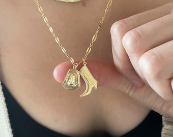 May include: A gold chain necklace with two charms: a cowboy hat and a cowboy boot. The charms are hanging from the chain and are visible in the image.