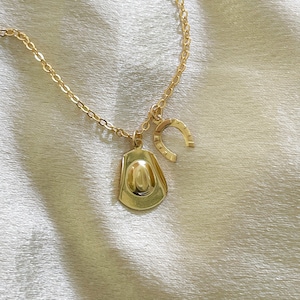 Op de afbeelding: Goudkleurige ketting met een cowboyhoed- en hoefijzerbedel. De ketting en bedels hebben een glanzende afwerking. De cowboyhoedbedel is gedetailleerd met een afgeronde kroon en rand. De hoefijzerbedel heeft een klassieke U-vorm.