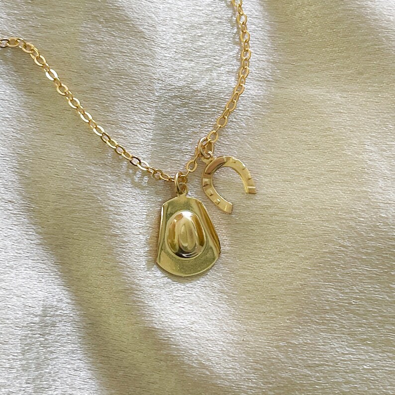 Op de afbeelding: Goudkleurige ketting met een cowboyhoed- en hoefijzerbedel. De ketting en bedels hebben een glanzende afwerking. De cowboyhoedbedel is gedetailleerd met een afgeronde kroon en rand. De hoefijzerbedel heeft een klassieke U-vorm.