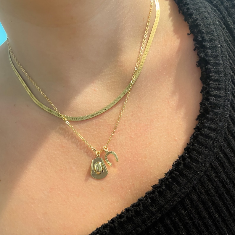 Op de afbeelding: Goudkleurige ketting set met twee kettingen. De bovenste ketting is een plat, slangenketting ontwerp. De onderste ketting heeft een cowboyhoed en een hoefijzer bedel. De sieraden worden getoond op een persoon die een zwarte top draagt.