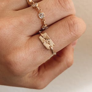 May include: Gold-toned ring featuring a rectangular charm with a starburst design. The ring has a delicate chain with clear, round gemstones. The ring is worn on a finger, showcasing the jewelry's intricate details and elegant design.