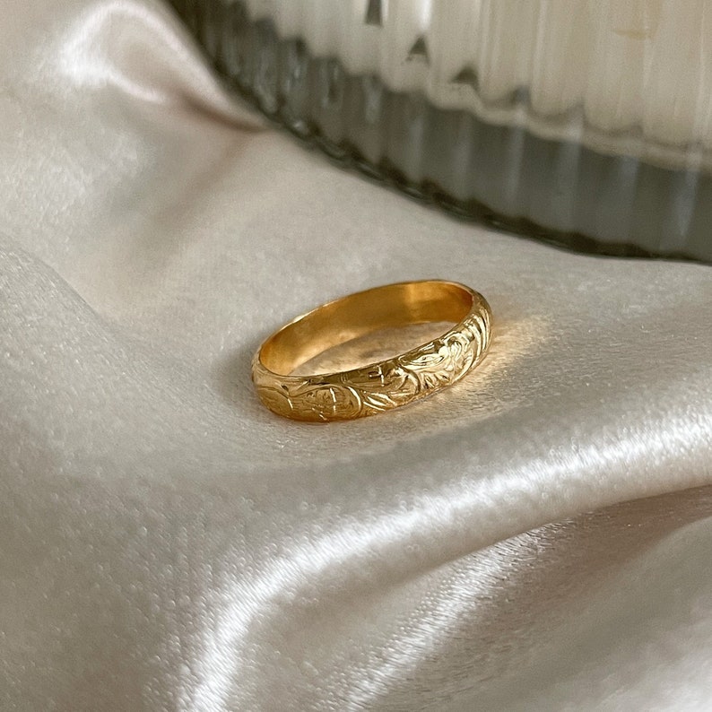 May include: A gold band ring with a floral design. The ring is on a white satin fabric.