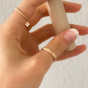 May include: A gold ring with a floral design on the band. The ring is worn on the thumb of a hand with a white nail polish manicure.