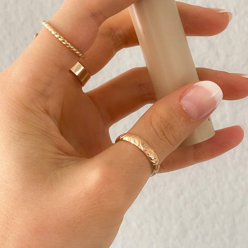 May include: A gold ring with a floral design on the band. The ring is worn on the thumb of a hand with a white nail polish manicure.