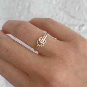 Könnte beinhalten: Ein zweifarbiger Ring mit Knoten-Design. Der Ring hat ein goldenes Band und ein silbernes Band, mit einem goldenen Knotendetail in der Mitte. Der Ring wird an einem Finger getragen.
