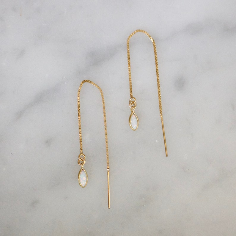 May include: A pair of gold-colored threader earrings with a white opal-like teardrop-shaped charm. The earrings have a delicate chain design and are displayed on a marble surface.