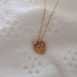 Puede incluir: Un collar de oro con un colgante en forma de corazón, un pequeño dije de corazón y un dije de cristal transparente. El collar está sobre un fondo blanco.