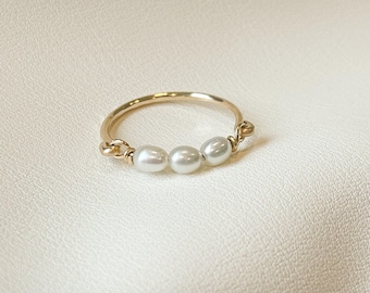 May include: A delicate gold ring featuring three white pearls. The pearls are evenly spaced across the top of the ring, which has a thin, polished band. The ring is set against a soft, off-white background.