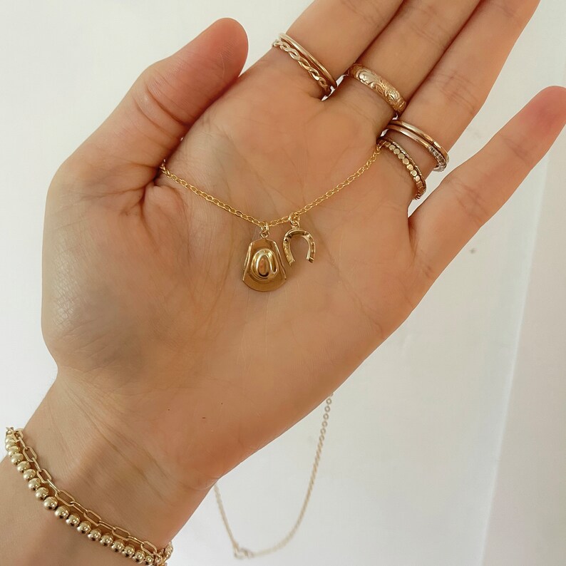 Op de afbeelding: Een goudkleurige ketting met een cowboyhoed- en hoefijzerhanger. De ketting wordt in een hand gehouden, en er zijn ook verschillende gouden ringen en een gouden armband zichtbaar. De sieraden hebben een glanzende afwerking.