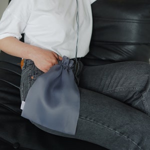 Puede incluir: Una mujer con una camiseta blanca y pantalones vaqueros azules está sentada en un sofá de cuero negro. Está sosteniendo una bolsa de cordón gris en su mano.