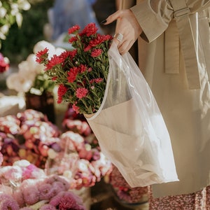 May include: A person holding a bouquet of pink and red flowers wrapped in a white mesh bag. The flowers are in a flower shop with other bouquets in the background.