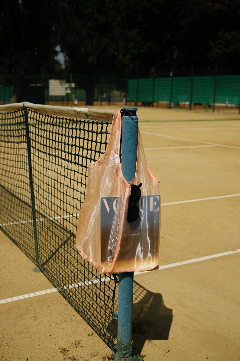 May include: A peach-colored mesh tote bag with a magazine inside. The magazine cover features the word "Vogue" in white letters. The bag is hanging on a blue metal pole on a tennis court.