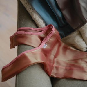 May include: A sheer pink fabric with a pink satin trim. The fabric has a subtle ombre effect, transitioning from a light pink to a darker pink. The fabric is folded and draped over a grey sofa.