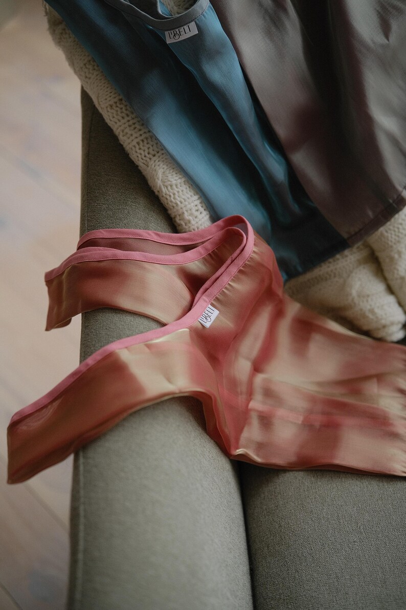 May include: A sheer pink fabric with a pink satin trim. The fabric has a subtle ombre effect, transitioning from a light pink to a darker pink. The fabric is folded and draped over a grey sofa.