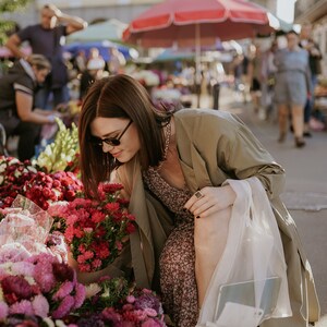 May include: A person browsing a flower market, surrounded by buckets of vibrant pink and red flowers. The person is wearing sunglasses, a floral dress, and a long olive-green coat. White boots and a gold chain are also visible.