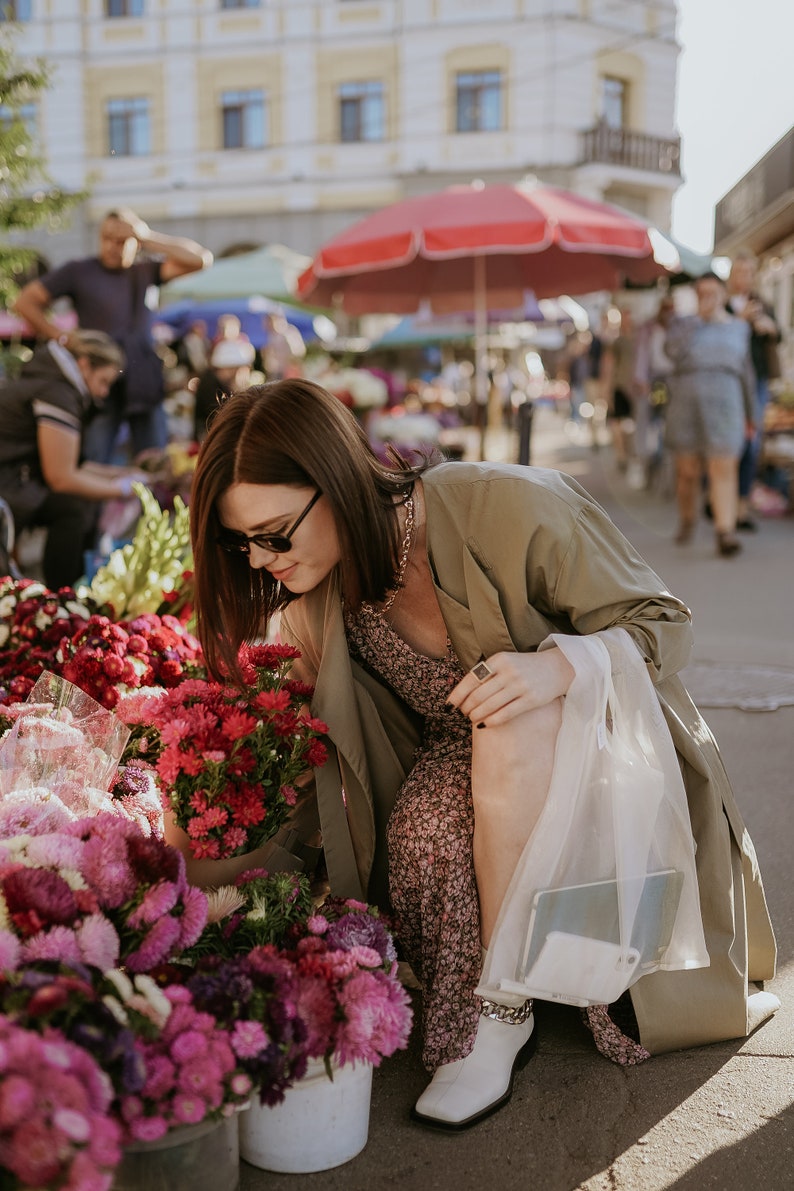 May include: A person browsing a flower market, surrounded by buckets of vibrant pink and red flowers. The person is wearing sunglasses, a floral dress, and a long olive-green coat. White boots and a gold chain are also visible.
