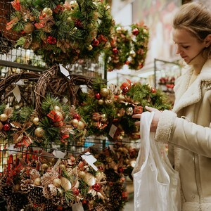 May include: Christmas wreaths on display, featuring green pine needles, gold ornaments, red bows, and pinecones. A woman in a cream-colored jacket holds a shopping bag, browsing the selection of festive holiday decorations.