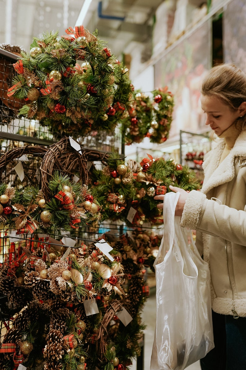 May include: Christmas wreaths on display, featuring green pine needles, gold ornaments, red bows, and pinecones. A woman in a cream-colored jacket holds a shopping bag, browsing the selection of festive holiday decorations.