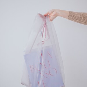 May include: A white mesh shopping bag with pink handles, holding a purple gift bag with the text "LA BOUSSOLE" in pink.