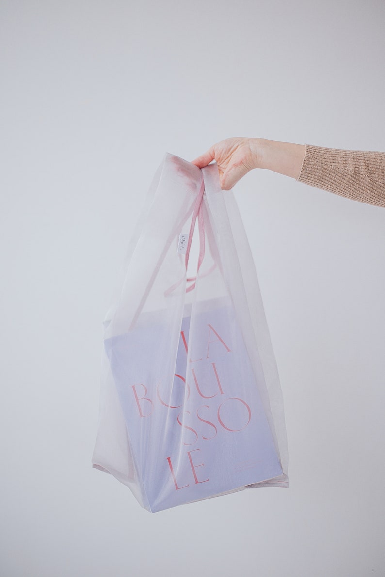 May include: A white mesh shopping bag with pink handles, holding a purple gift bag with the text "LA BOUSSOLE" in pink.
