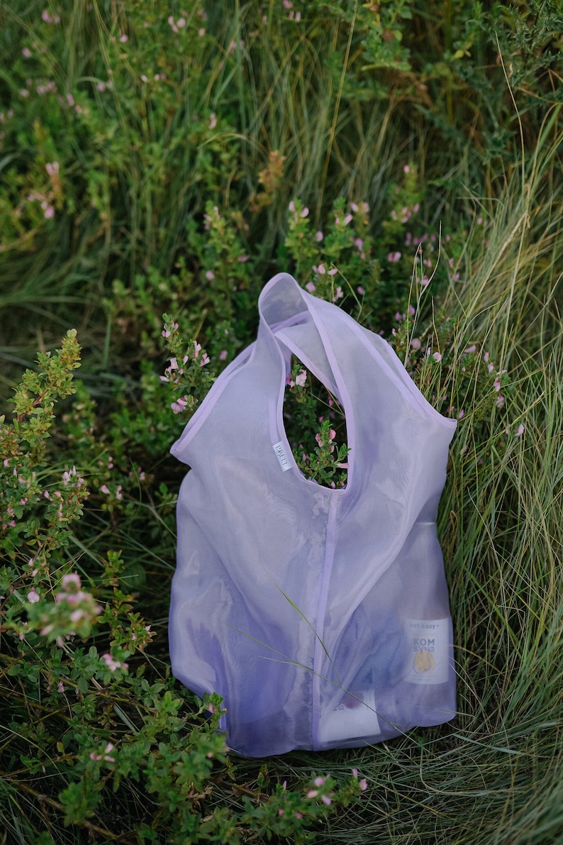 May include: A light purple reusable mesh shopping bag with the word "LILLA" printed on the side. The bag is lying on a bed of green grass and pink flowers.