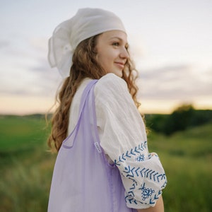 May include: A woman wearing a white headscarf and a white embroidered blouse with blue stitching, holds a lavender reusable shopping bag. She is standing in a field of green grass.