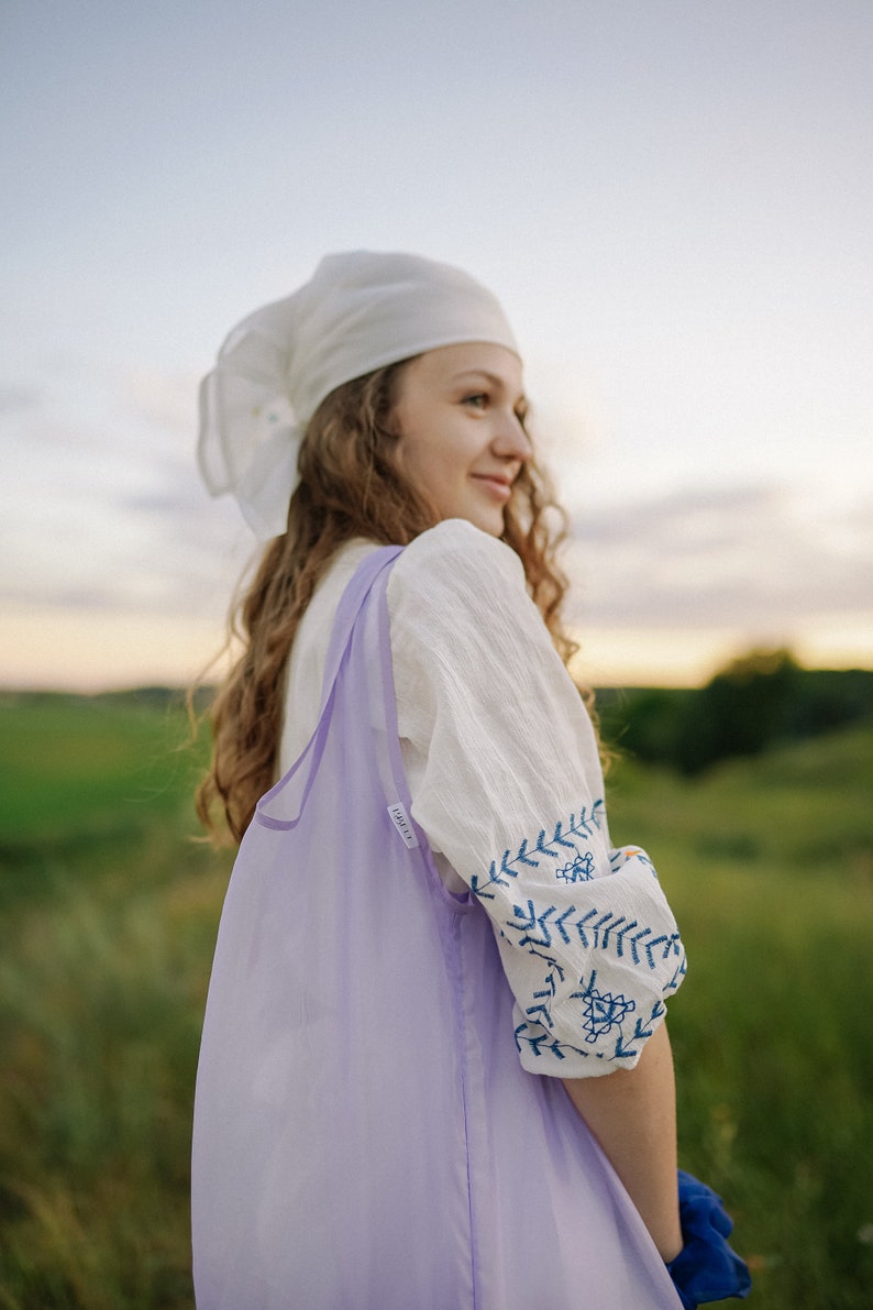 May include: A woman wearing a white headscarf and a white embroidered blouse with blue stitching, holds a lavender reusable shopping bag. She is standing in a field of green grass.