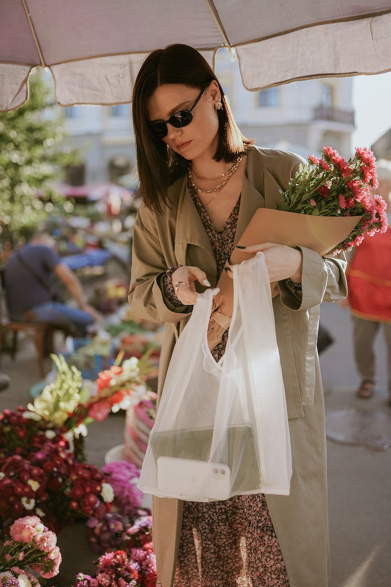 Puede incluir: Una mujer con un abrigo beige de gabardina sostiene un ramo de flores rosas envuelto en papel marr&oacute;n. Lleva una bolsa de la compra reutilizable blanca en la otra mano. Las flores est&aacute;n enfocadas, y la mujer est&aacute; desenfocada.