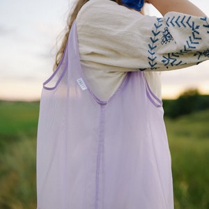 May include: A woman wearing a white embroidered top and a blue scrunchie carries a lavender mesh tote bag with two bottles of kombucha inside. The bottles have a label that reads "eat easy" and "kombucha".