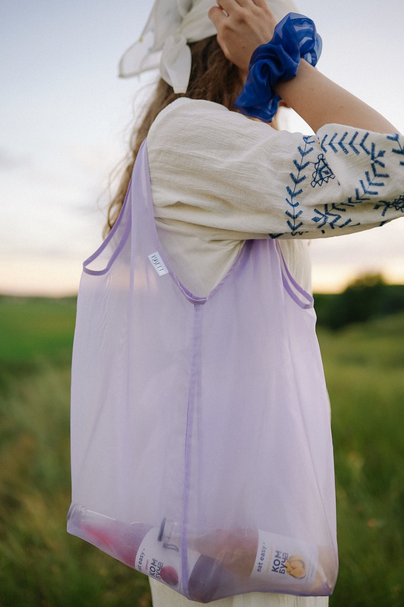 May include: A woman wearing a white embroidered top and a blue scrunchie carries a lavender mesh tote bag with two bottles of kombucha inside. The bottles have a label that reads "eat easy" and "kombucha".