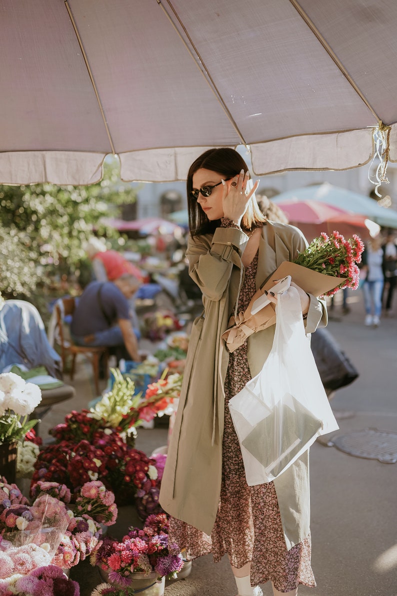 Puede incluir: Una mujer con un abrigo beige y un vestido floral est&aacute; comprando flores en un mercado. Lleva un ramo de flores rosas y una bolsa de pl&aacute;stico blanca.