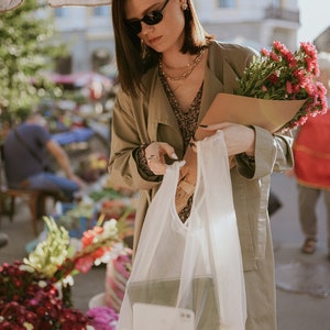 May include: A woman in sunglasses and a long coat holds a bouquet of red flowers wrapped in brown paper. She also holds a white mesh bag containing a tablet and a smartphone. The scene takes place at an outdoor flower market.