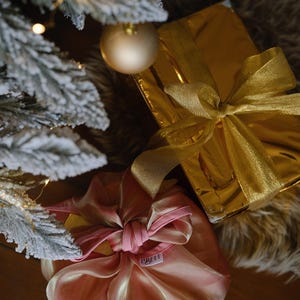May include: Two Christmas presents wrapped in gold and pink paper with matching ribbons. The gold gift is wrapped in shiny paper with a gold ribbon. The pink gift is wrapped in a sheer pink fabric with a pink ribbon. The presents are sitting on a brown wooden surface.