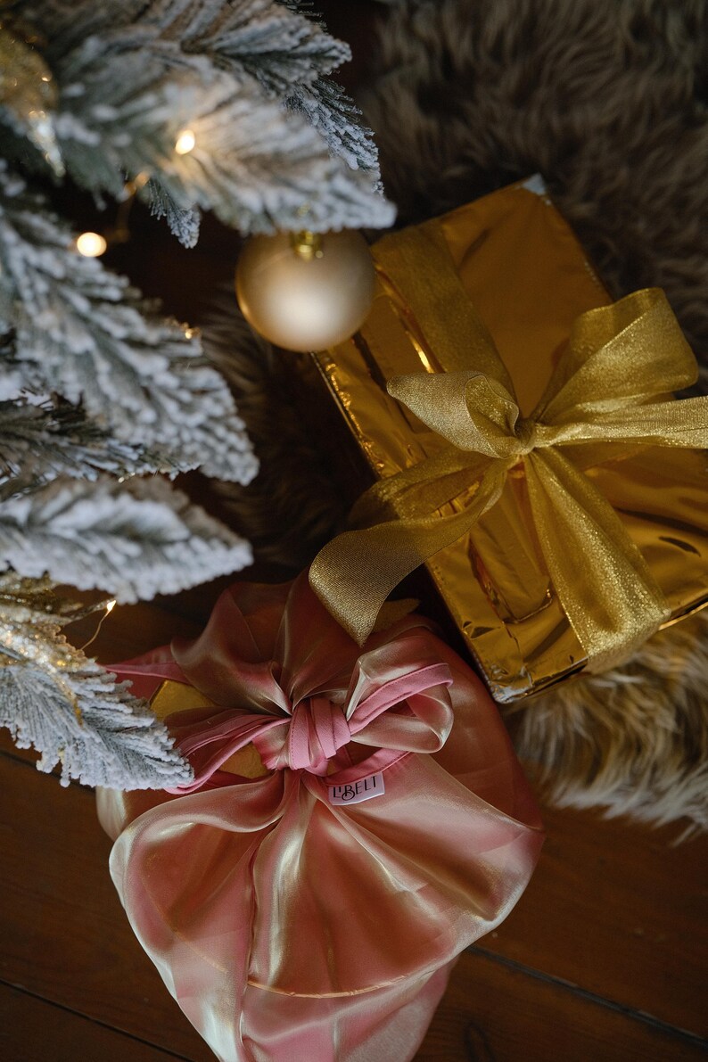 May include: Two Christmas presents wrapped in gold and pink paper with matching ribbons. The gold gift is wrapped in shiny paper with a gold ribbon. The pink gift is wrapped in a sheer pink fabric with a pink ribbon. The presents are sitting on a brown wooden surface.