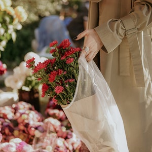 Puede incluir: Un ramo de flores rosas y rojas envuelto en una bolsa de papel y pl&aacute;stico blanca. Las flores las sostiene una persona que lleva una gabardina beige.