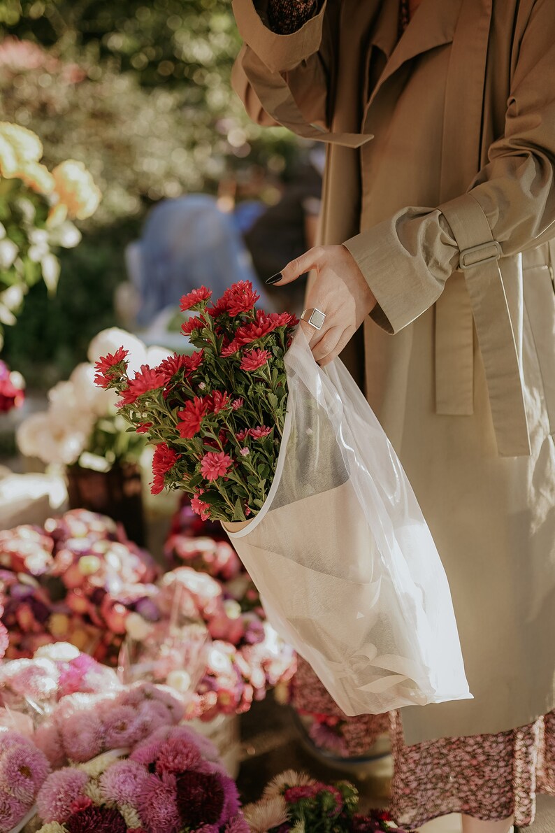 Puede incluir: Un ramo de flores rosas y rojas envuelto en una bolsa de papel y pl&aacute;stico blanca. Las flores las sostiene una persona que lleva una gabardina beige.