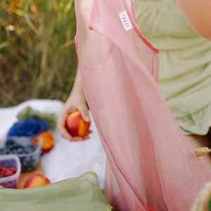 May include: A pink sheer sleeveless top with a white label that says "L'&Eacute;CH&Eacute;ANCE". The top is being held up by a person in a green dress. The person is sitting on a white blanket with fruit and a green bag.