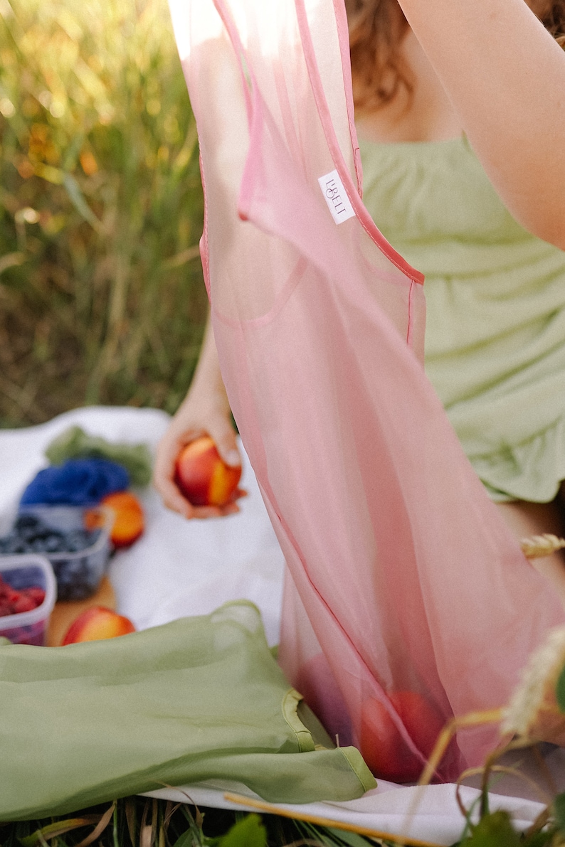 May include: A pink sheer sleeveless top with a white label that says "L'&Eacute;CH&Eacute;ANCE". The top is being held up by a person in a green dress. The person is sitting on a white blanket with fruit and a green bag.