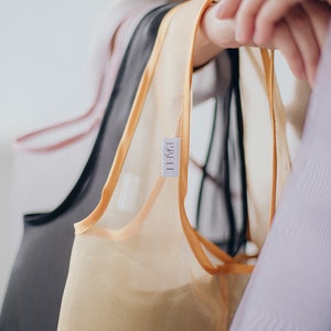 May include: A set of four sheer tops in different colors, including black, beige, pink, and purple. The tops have a delicate, silky texture and are trimmed with a thin, gold ribbon. The tops are hanging on a rack and have a label that reads "LABEL."