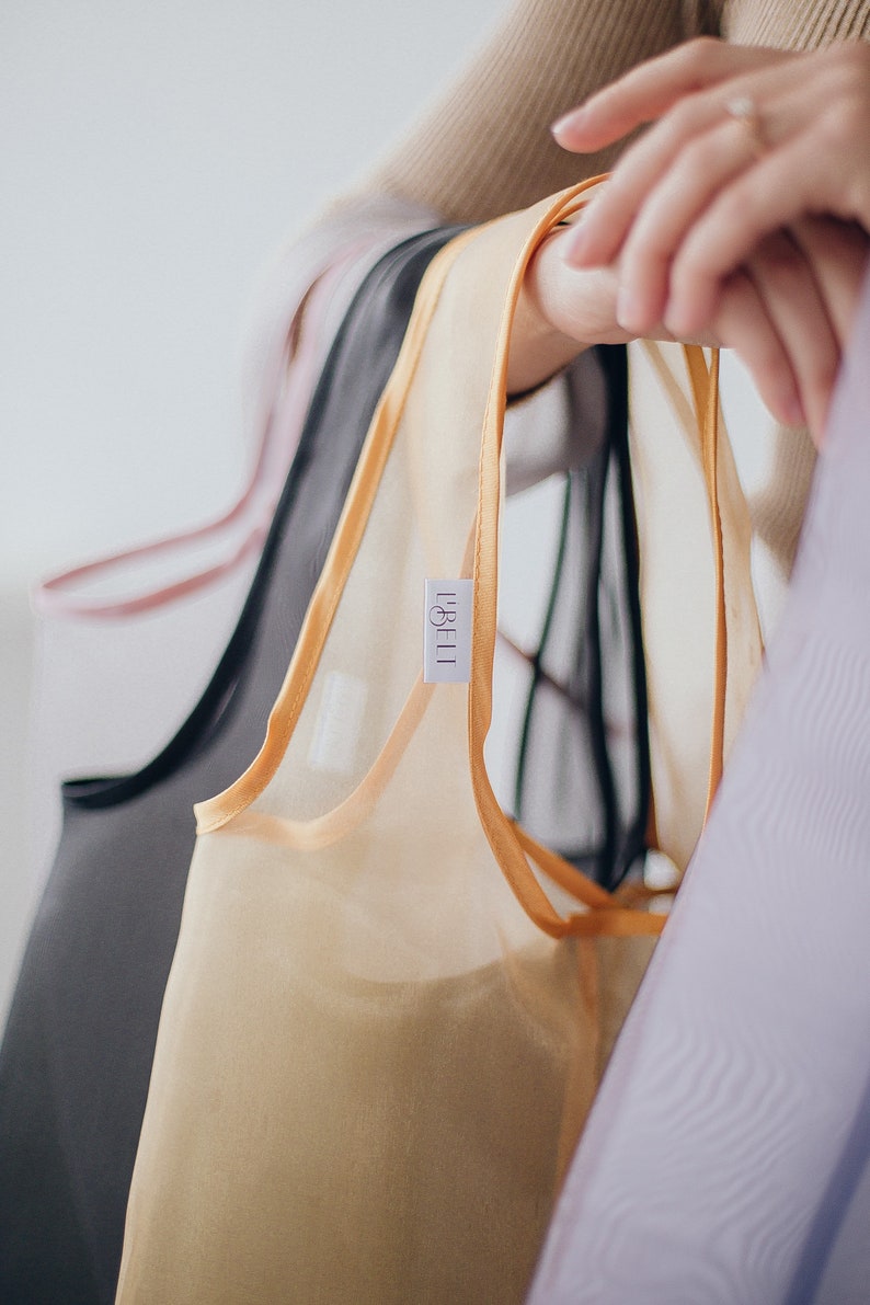May include: A set of four sheer tops in different colors, including black, beige, pink, and purple. The tops have a delicate, silky texture and are trimmed with a thin, gold ribbon. The tops are hanging on a rack and have a label that reads "LABEL."