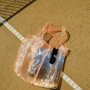 May include: A sheer pink mesh tote bag with a magazine and sunglasses inside. The bag is lying on a brown surface with a white line.